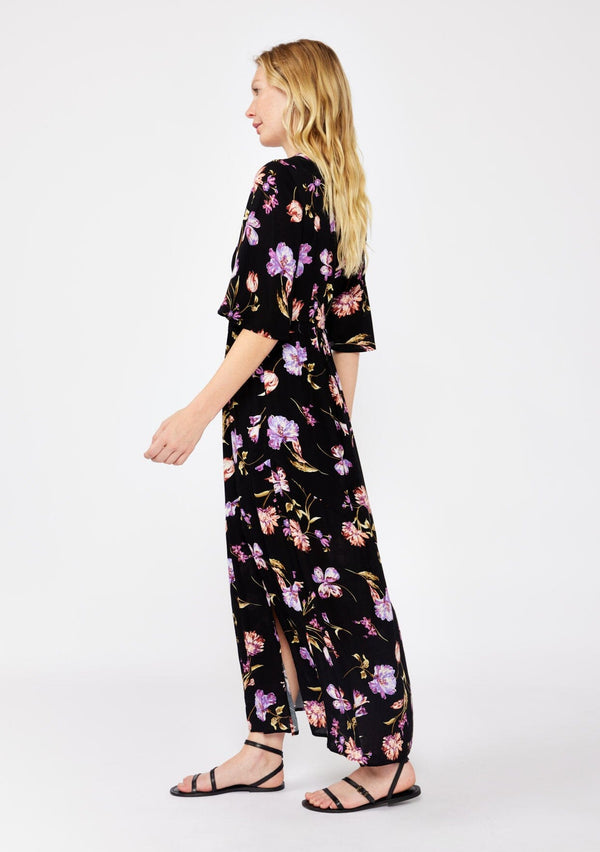 [Color: Black/Purple] A blonde model wearing a classic bohemian silhouette in a black and purple floral print. A relaxed fit maxi dress with flowy short sleeves, plunging v neckline, empire waistline with a front elastic waist, and side slit. Styled with casual black strappy sandals for a day to night look.