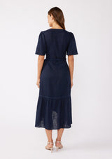 [Color: Navy] A back facing image of a brunette model wearing a navy bohemian mid length wrap dress. With short flutter sleeves, embroidered detail, a deep v neckline, a side tie waist closure, and a flowy tiered skirt.