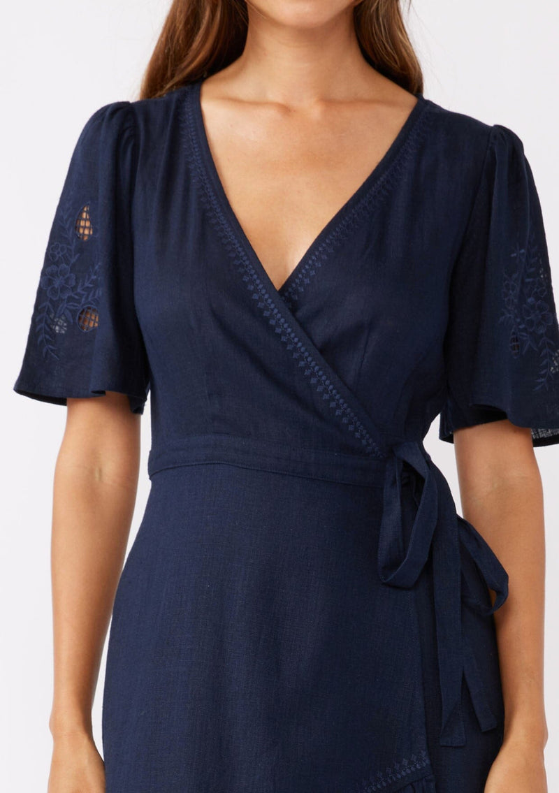 [Color: Navy] A detailed shot of a brunette model wearing a navy bohemian mid length wrap dress. With short flutter sleeves, embroidered detail, a deep v neckline, a side tie waist closure, and a flowy tiered skirt.