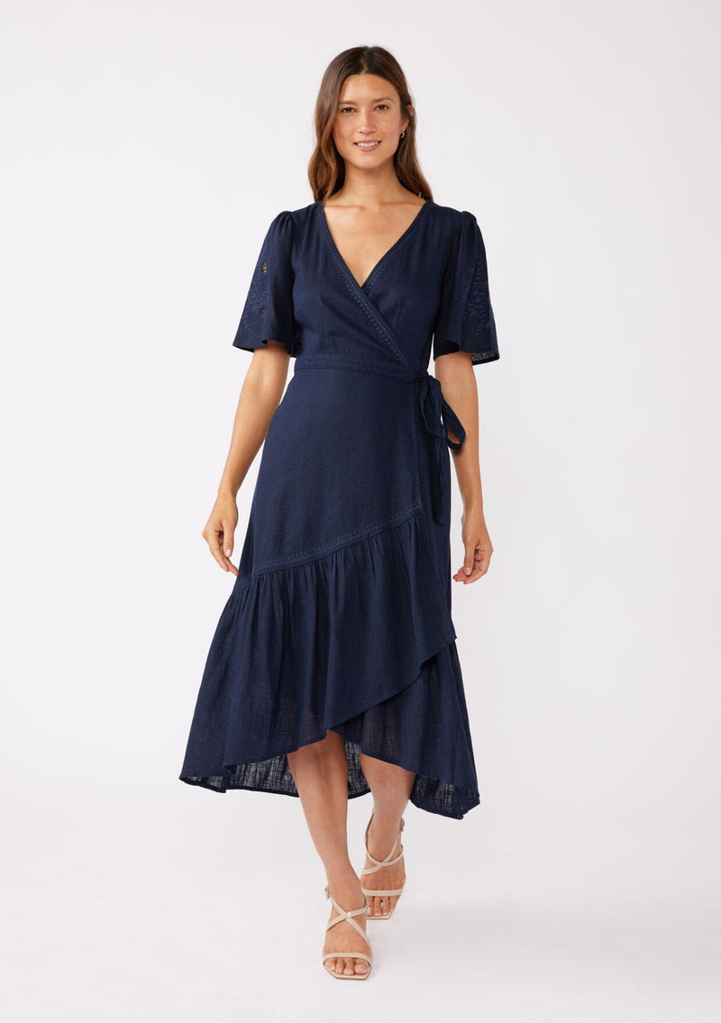 [Color: Navy] A front facing image of a brunette model wearing a navy bohemian mid length wrap dress. With short flutter sleeves, embroidered detail, a deep v neckline, a side tie waist closure, and a flowy tiered skirt.