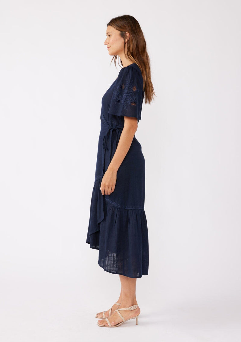 [Color: Navy] A side facing image of a brunette model wearing a navy bohemian mid length wrap dress. With short flutter sleeves, embroidered detail, a deep v neckline, a side tie waist closure, and a flowy tiered skirt.