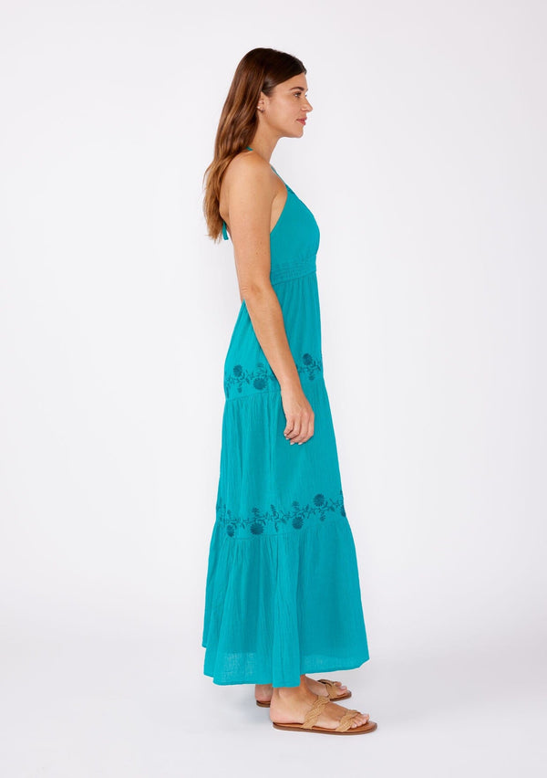 [Color: Teal Blue] A brunette model wearing a teal bohemian maxi dress. A classic halter maxi dress with a v neck, smocked empire waist, tiered skirt, and floral embroidered details.