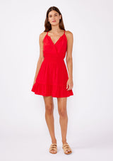 [Color: Poppy Red] A brunette woman wearing a casual bright red mini dress with a sleeveless design, split v neckline, smocked waistline, and a ruffle trimmed hem. A casual yet formal dress perfect for date night and special occasions.