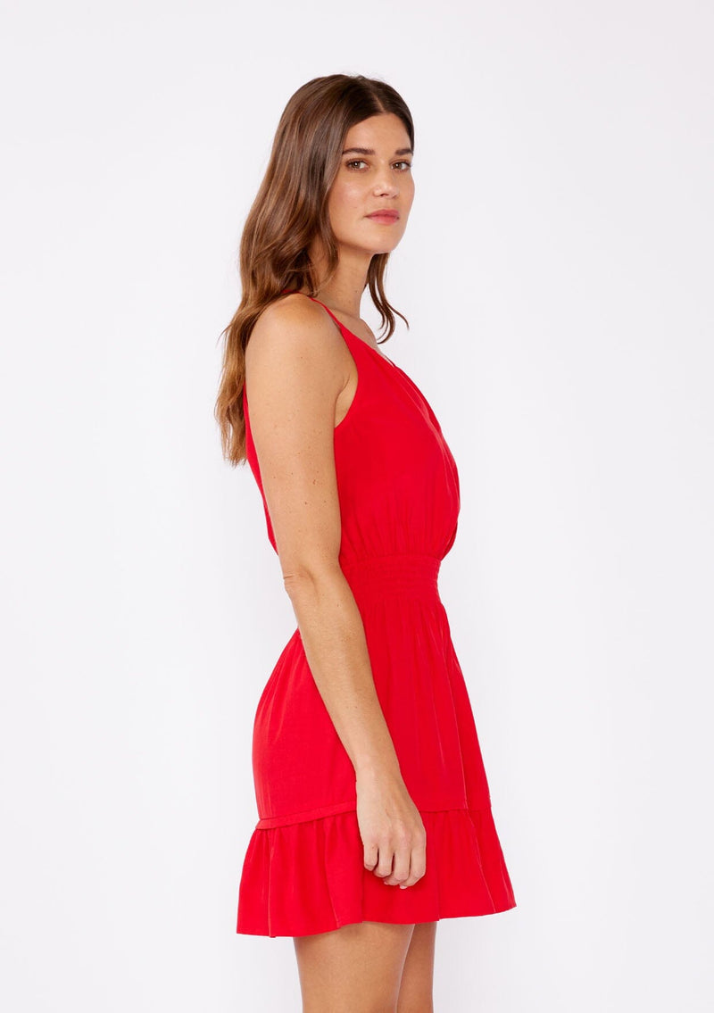 [Color: Poppy Red] A brunette woman wearing a casual bright red mini dress with a sleeveless design, split v neckline, smocked waistline, and a ruffle trimmed hem. A casual yet formal dress perfect for date night and special occasions.