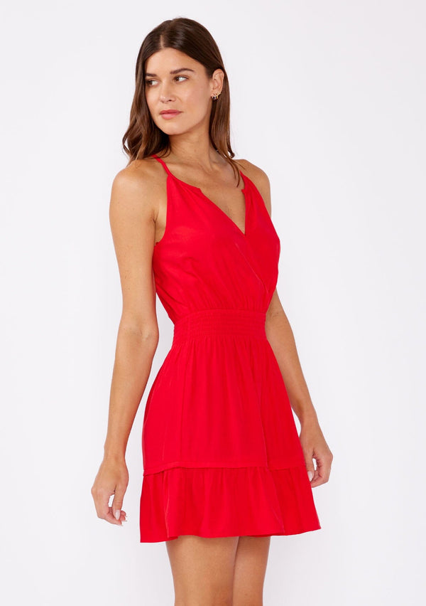 [Color: Poppy Red] A brunette woman wearing a casual bright red mini dress  with a sleeveless design, split v neckline, smocked waistline, and a ruffle trimmed hem. A casual yet formal dress perfect  for date night and special occasions. 