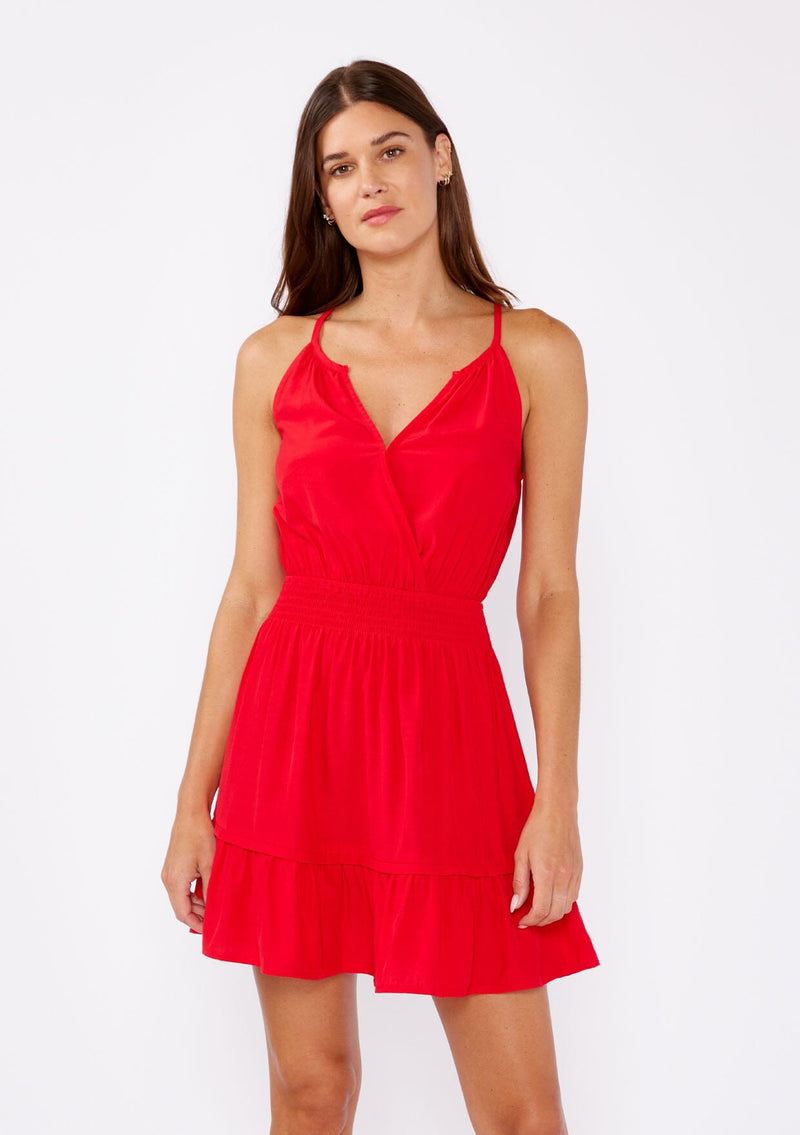 [Color: Poppy Red] A brunette woman wearing a casual bright red mini dress with a sleeveless design, split v neckline, smocked waistline, and a ruffle trimmed hem. A casual yet formal dress perfect for date night and special occasions.