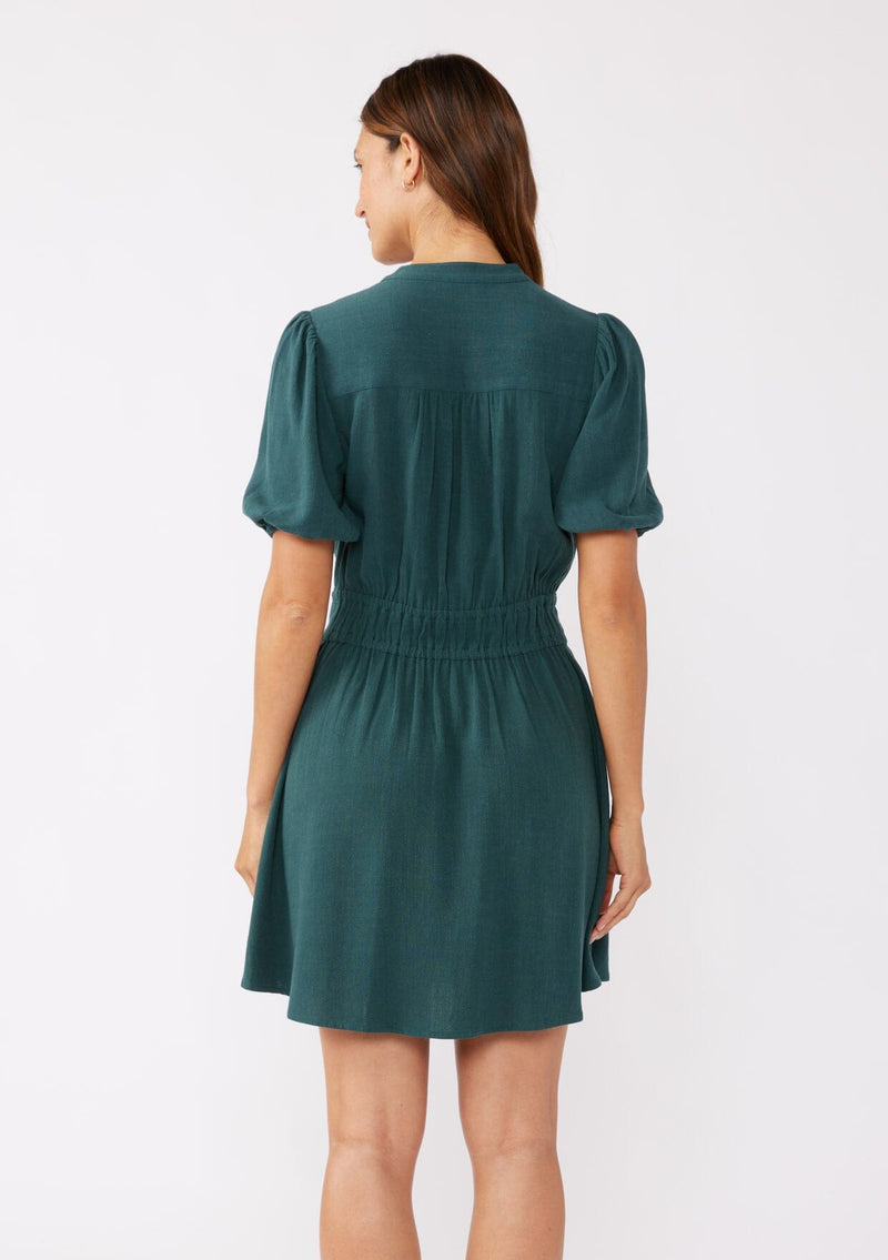 A back image of a brunette model wearing a classic Fall bohemian mini dress in a green teal. Crafted with a rayon and linen blend, this modern button front mini dress features short puff sleeves, a button front, and two adjustable drawstrings. 