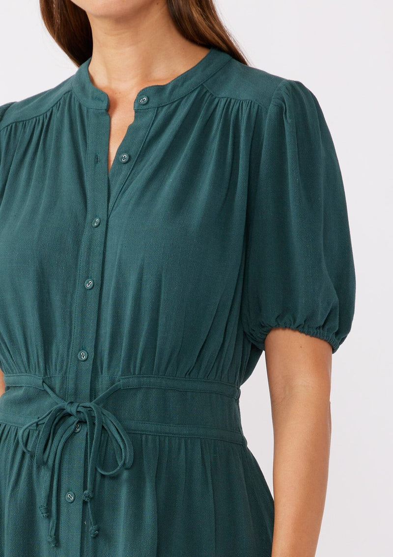 A detailed image of a brunette model wearing a classic Fall bohemian mini dress in a green teal. Crafted with a rayon and linen blend, this modern button front mini dress features short puff sleeves, a button front, and two adjustable drawstrings. 