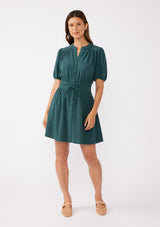 [Color: Dusty Teal] A front image of a brunette model wearing a classic bohemian mini dress in a green teal. Crafted with a rayon and linen blend, this modern button front mini dress features short puff sleeves, a button front, and two adjustable drawstrings. Styled with ballet flats for cool and casual look for the Fall season. 