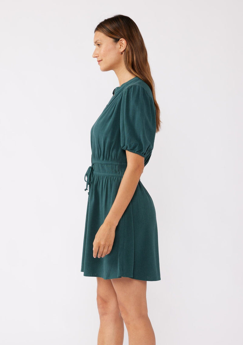 A side image of a brunette model wearing a classic Fall bohemian mini dress in a green teal. Crafted with a rayon and linen blend, this modern button front mini dress features short puff sleeves, a button front, and two adjustable drawstrings. 