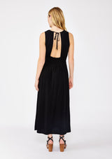 [Color: Black] A blonde model wearing a sophisticated modern bohemian midi dress with a shirred bodice. This classic black silhouette features a sleeveless design, round neckline, side pockets, and a flowy skirt. Completed with a sexy open back with tie detail and styled with comfortable flat sandals for laid back look.