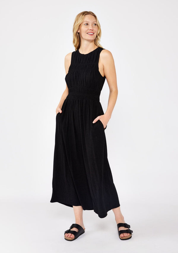 [Color: Black] A blonde model wearing a sophisticated modern bohemian midi dress with a shirred bodice. This classic black silhouette features a sleeveless design, round neckline, side pockets, and a flowy skirt. Completed with a sexy open back with tie detail and styled with comfortable flat sandals for laid back look.