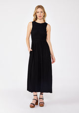 [Color: Black] A blonde model wearing a sophisticated modern bohemian midi dress with a shirred bodice. This classic black silhouette features a sleeveless design, round neckline, side pockets, and a flowy skirt. Completed with a sexy open back with tie detail and styled with comfortable flat sandals for laid back look.