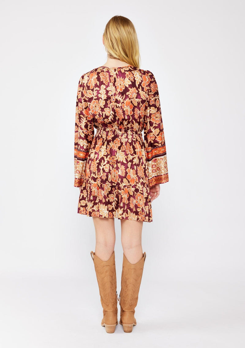 [Color: Wine/Dusty Rose] A blonde model wearing an ultra bohemian mini dress in a burgundy and orange. A floral border print dress with long bell sleeves, smocked waistline, split v neckline, and flowy skirt. Styled with camel knee length boots for the fall season. 