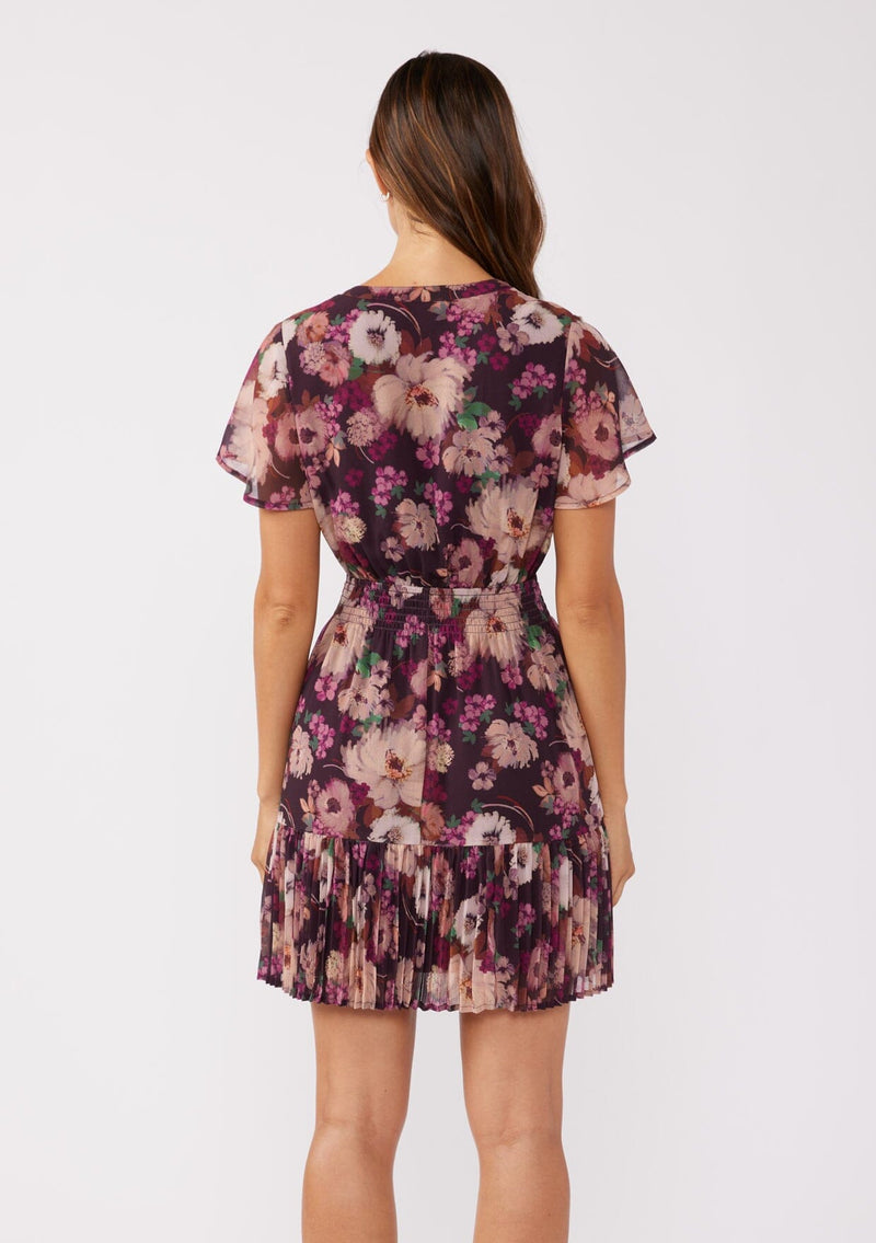 [Color: Plum/Dusty Rose] A back facing image of a brunette model wearing a flowy chiffon mini dress in a purple floral pattern. The dress showcases a split v-neckline, fluttery short sleeves, a smocked waist, and a pleated skirt. Paired with black formal heels, it's a perfect choice for fall, the holiday season, and special occasions.