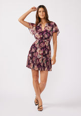 [Color: Plum/Dusty Rose] Front-facing full-body image of a brunette model wearing a flowy chiffon mini dress in a purple floral pattern. The dress showcases a split v-neckline, fluttery short sleeves, a smocked waist, and a pleated skirt. Paired with black formal heels, it's a perfect choice for fall, the holiday season, and special occasions.