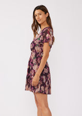 [Color: Plum/Dusty Rose] A side facing image of a brunette model wearing a flowy chiffon mini dress in a purple floral pattern. The dress showcases a split v-neckline, fluttery short sleeves, a smocked waist, and a pleated skirt. Paired with black formal heels, it's a perfect choice for fall, the holiday season, and special occasions.