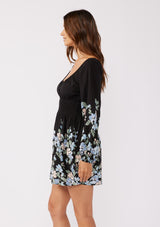 [Color: Black/Pale Blue] A side facing image of a brunette model wearing a black mini dress adorned with a pale blue floral print. The dress features a scoop neckline, long sleeves, and a smocked waistline. Styled with ballet flats, the fit-and-flare design offers a chic look for any special occasion.