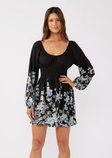 [Color: Black/Pale Blue] A front facing image of a brunette model wearing a black mini dress adorned with a pale blue floral print. The dress features a scoop neckline, long sleeves, and a smocked waistline. Styled with ballet flats, the fit-and-flare design offers a chic look for any special occasion.