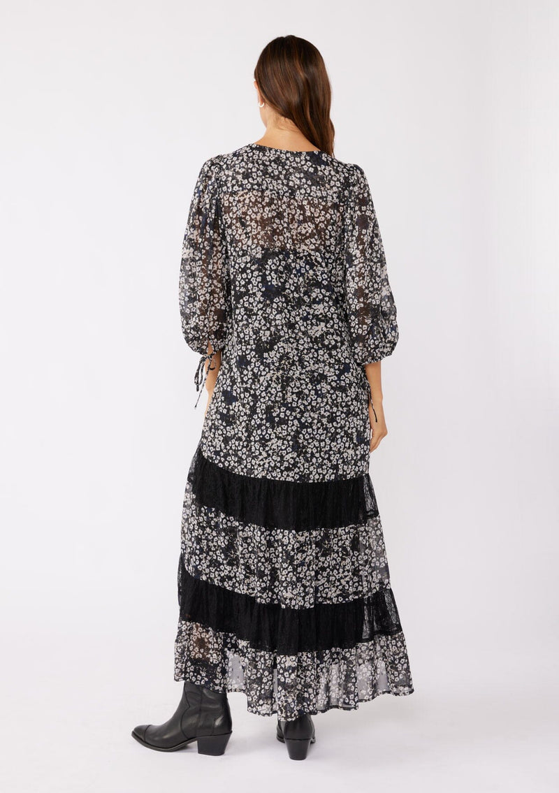 [Color: Black/Grey] A brunette model wearing a black floral print maxi , crafted in an ultra-relaxed fit for the fall season. With a split v neckline and tassel ties, volume sleeves, and a flowy tiered skirt with lace trim inserts. Styled with black boots for the ultimate fall look.