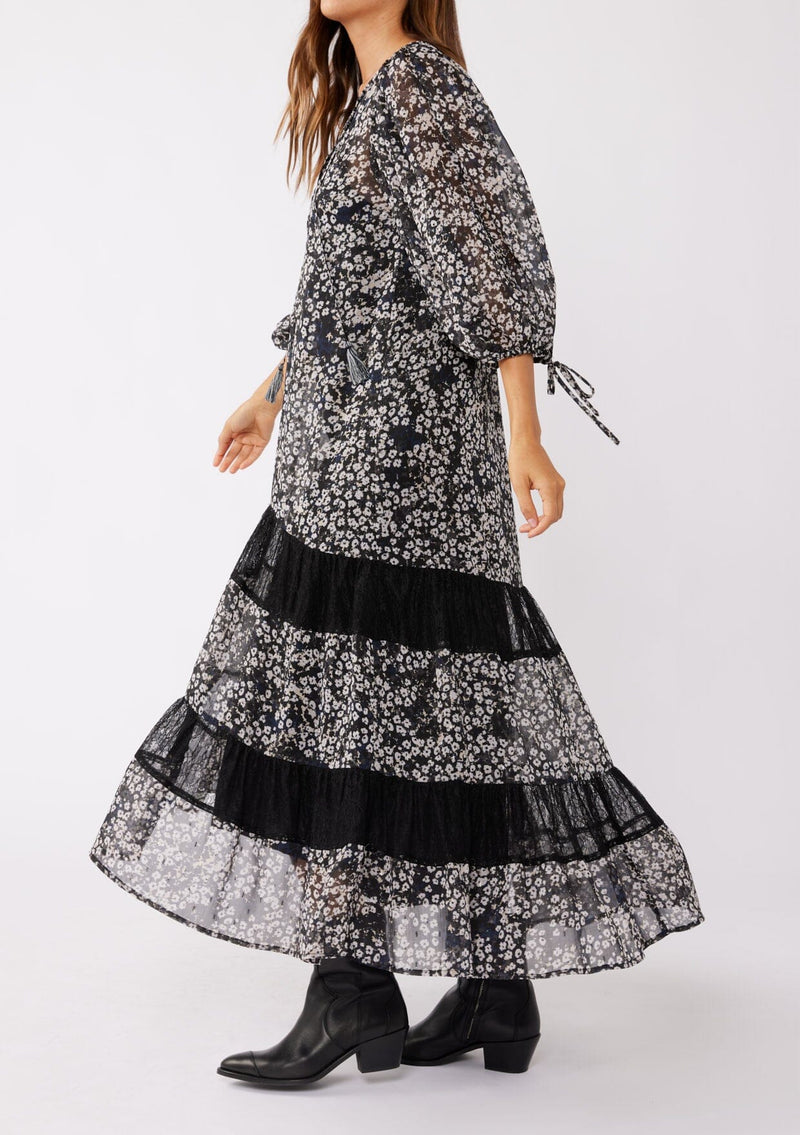 [Color: Black/Grey] A brunette model wearing a black floral print maxi , crafted in an ultra-relaxed fit for the fall season. With a split v neckline and tassel ties, volume sleeves, and a flowy tiered skirt with lace trim inserts. Styled with black boots and a hat for the ultimate fall look.