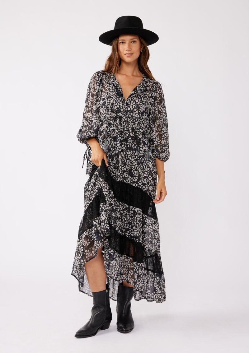 [Color: Black/Grey] A brunette model wearing a black floral print maxi , crafted in an ultra-relaxed fit for the fall season. With a split v neckline and tassel ties, volume sleeves, and a flowy tiered skirt with lace trim inserts. Styled with black boots and a hat for the ultimate fall look.