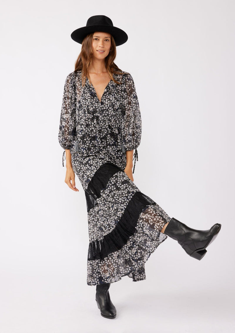 [Color: Black/Grey] A brunette model wearing a black floral print maxi , crafted in an ultra-relaxed fit for the fall season. With a split v neckline and tassel ties, volume sleeves, and a flowy tiered skirt with lace trim inserts. Styled with black boots and a hat for the ultimate fall look.