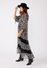 [Color: Black/Grey] A brunette model wearing a black floral print maxi , crafted in an ultra-relaxed fit for the fall season. With a split v neckline and tassel ties, volume sleeves, and a flowy tiered skirt with lace trim inserts. Styled with black boots and a hat for the ultimate fall look.