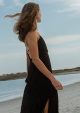 [Color: Black] A side image of a brunette model wearing a sleeveless, pleated maxi dress. This special occasion dress features a surplice v neckline, empire waistline, an adjustable strappy back, and a side slit. Perfect for the holidays or any special event. 