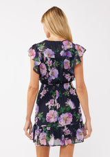 [Color: Navy/Lilac] A back-facing image of a blonde model wearing a navy and purple floral mini dress. The dress features lace detailing at the shoulder, a surplice v-neckline, a smocked waist, and a flowy tiered skirt. Ideal for dinners, special occasions, and vacation looks.