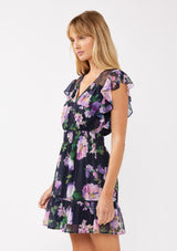 [Color: Navy/Lilac] A side-facing image of a blonde model wearing a navy and purple floral mini dress. The dress features lace detailing at the shoulder, a surplice v-neckline, a smocked waist, and a flowy tiered skirt. Ideal for dinners, special occasions, and vacation looks.