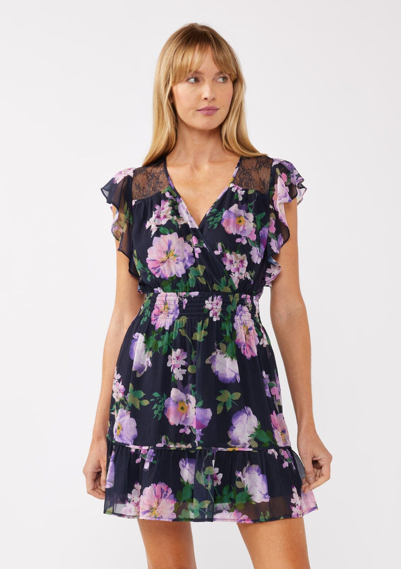 [Color: Navy/Lilac] A front-facing image of a blonde model wearing a navy and purple floral mini dress. The dress features lace detailing at the shoulder, a surplice v-neckline, a smocked waist, and a flowy tiered skirt. Ideal for dinners, special occasions, and vacation looks.