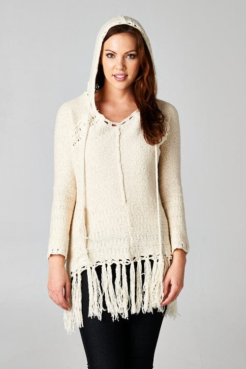 [Color: Natural] A brunette model wearing an off white vintage bohemian knit fringed sweater with a tie string hood and a long hemline. 