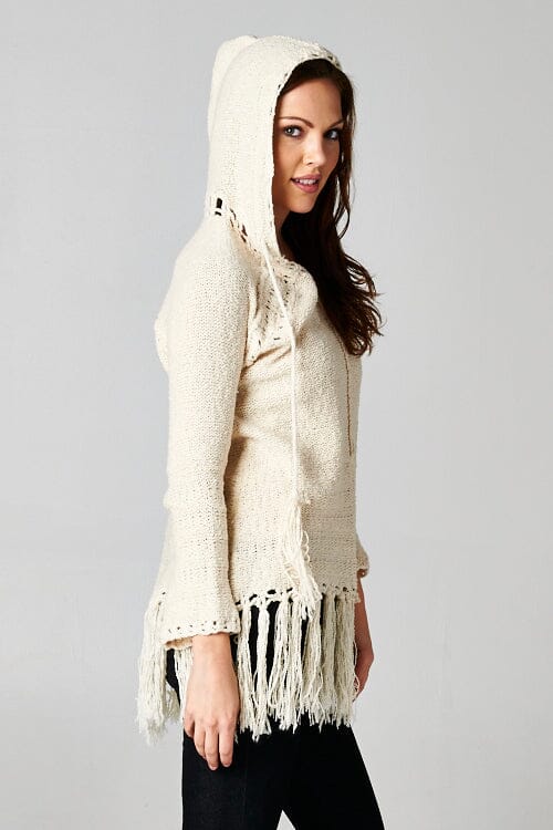 [Color: Natural] A brunette model wearing an off white vintage bohemian knit fringed sweater with a tie string hood and a long hemline. 