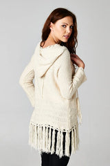 [Color: Natural] A brunette model wearing an off white vintage bohemian knit fringed sweater with a tie string hood and a long hemline. 