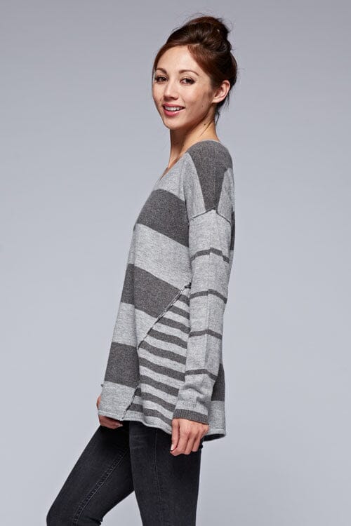 [Color: Charcoal/Heather Grey] A side facing image of a brunette model wearing a grey patchwork striped knit pullover with a v-neckline, long sleeves, dropped shoulders, and exposed seams. 