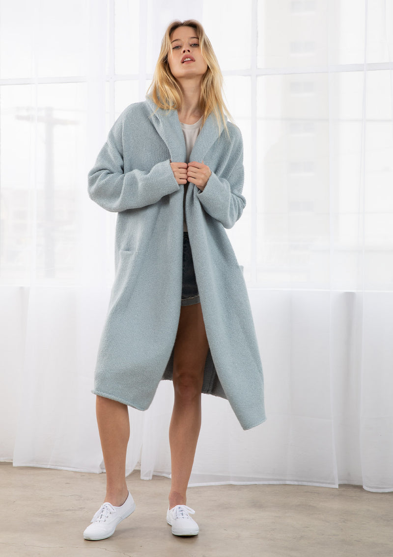 [Color: Ice Blue] A full body front facing image of a blonde model wearing a best selling oversized cardigan in a blue color. A cozy and thick sweater coat with a hood, an open front, side pockets, a cocoon style silhouette, and a mid length hem. 