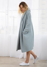 [Color: Ice Blue] A full body front facing image of a blonde model wearing a best selling oversized cardigan in a blue color. A cozy and thick sweater coat with a hood, an open front, side pockets, a cocoon style silhouette, and a mid length hem. 