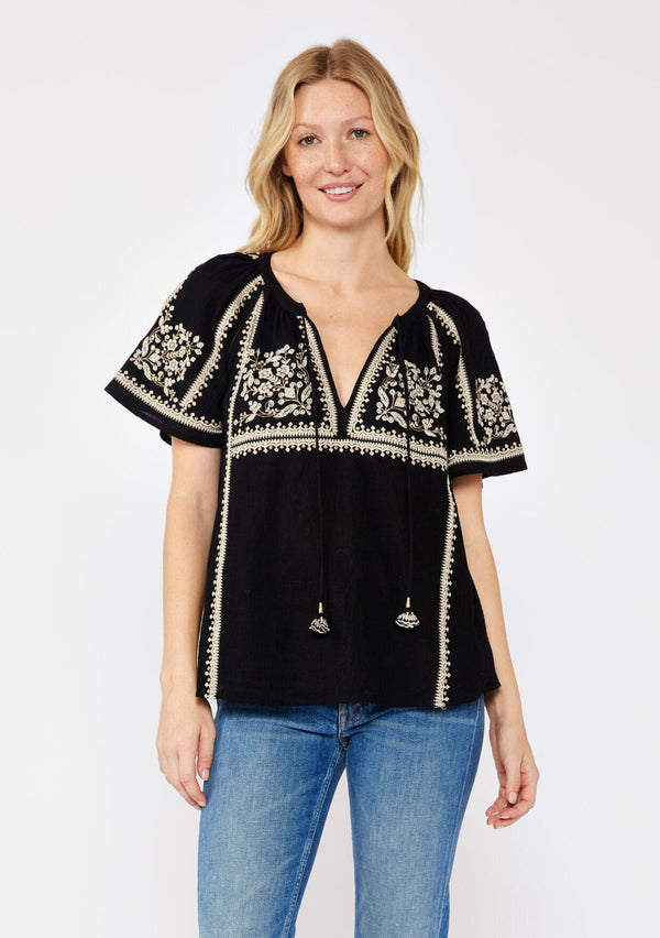 [Color: Black/Natural] A front facing image of a blonde model wearing a black cotton top with bohemian beige embroidered detail. With short raglan sleeves, a split v neckline with ties, and a relaxed fit.