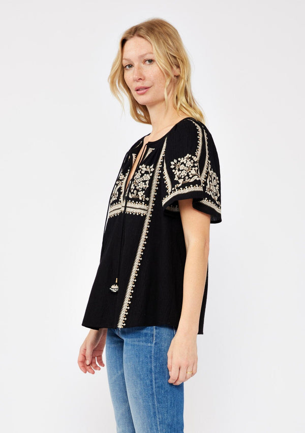 [Color: Black/Natural] A side facing image of a blonde model wearing a black cotton top with bohemian beige embroidered detail. With short raglan sleeves, a split v neckline with ties, and a relaxed fit.