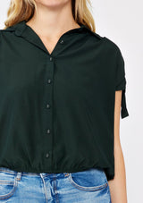 [Color: Olive] A blonde model wearing a dark green olive cropped top. With a collared neckline, button front, ruched shoulder with adjustable ties, and an elastic hem. A casual fall top paired with denim jeans.