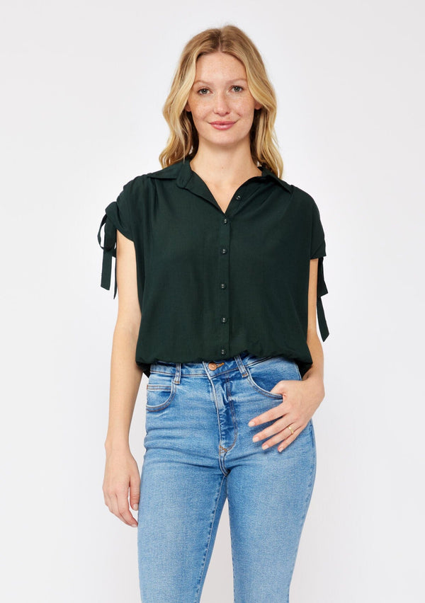 [Color: Olive] A blonde model wearing a dark green olive cropped top. With a collared neckline, button front, ruched shoulder with adjustable ties, and an elastic hem. A casual fall top paired with denim jeans. 
