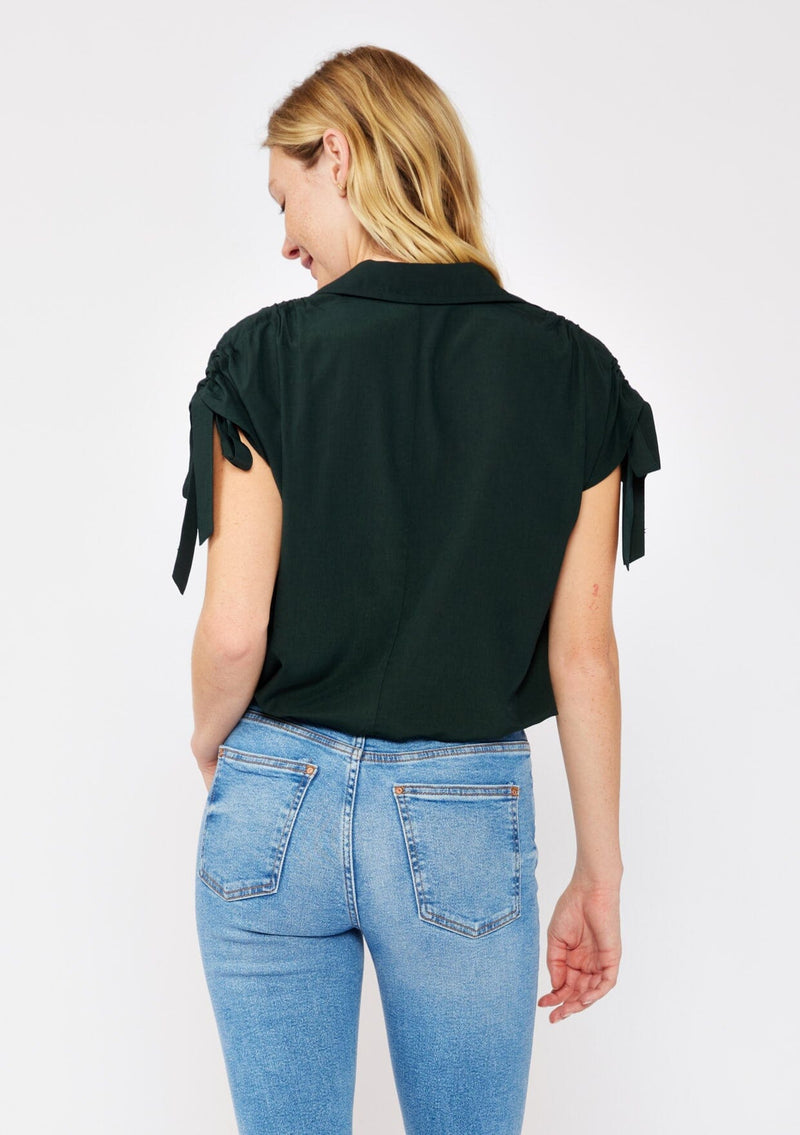 [Color: Olive] A blonde model wearing a dark green olive cropped top. With a collared neckline, button front, ruched shoulder with adjustable ties, and an elastic hem. A casual fall top paired with denim jeans.