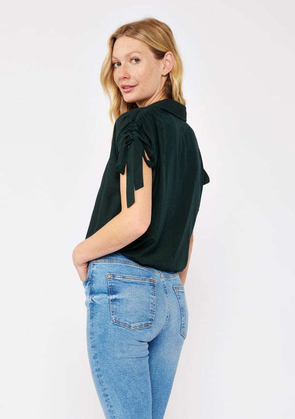 [Color: Olive] A blonde model wearing a dark green olive cropped top. With a collared neckline, button front, ruched shoulder with adjustable ties, and an elastic hem. A casual fall top paired with denim jeans.