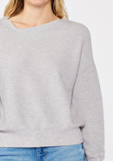 [Color: Heather Grey] A blonde model wearing a grey relaxed fit sweater with an asymmetrical neckline and long sleeves with a dropped shoulder. An everyday pullover sweater styled with denim jeans for the fall season.