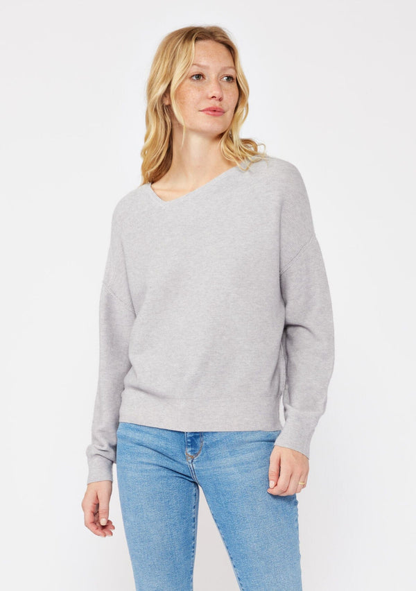 [Color: Heather Grey] A blonde model wearing a grey relaxed fit sweater with an asymmetrical neckline and long sleeves with a dropped shoulder. An everyday pullover sweater styled with denim jeans for the fall season. 