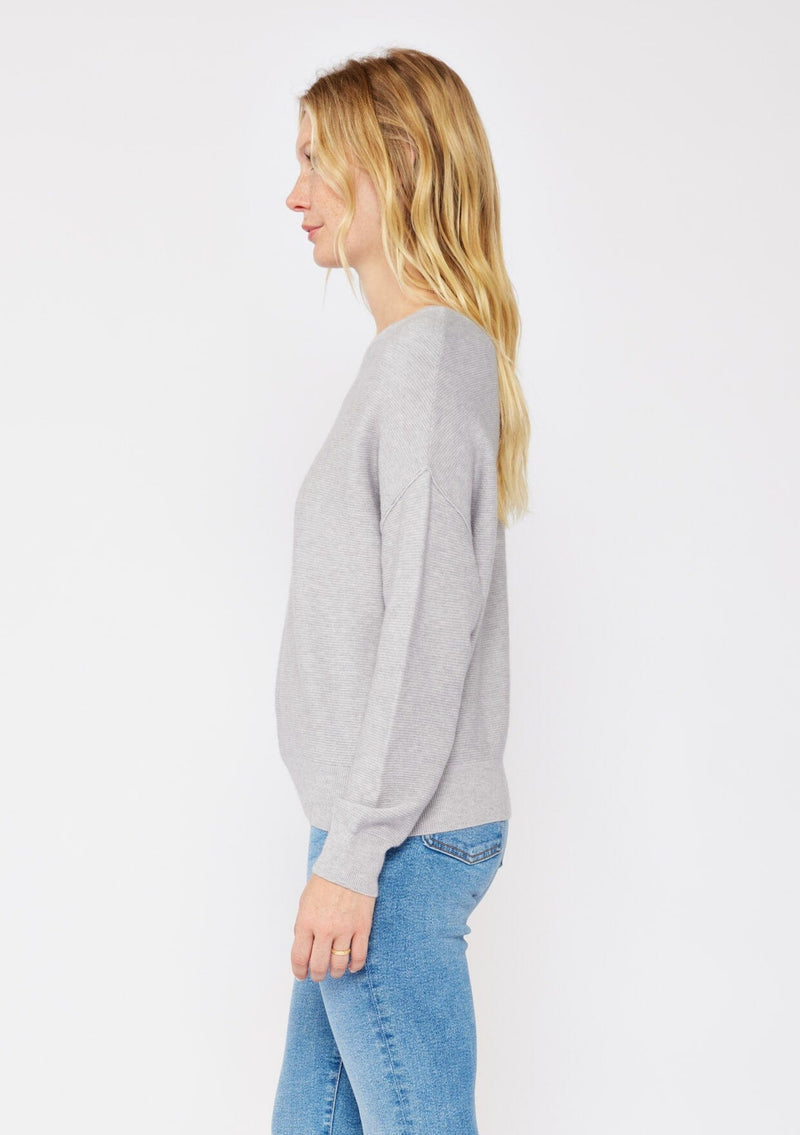 [Color: Heather Grey] A blonde model wearing a grey relaxed fit sweater with an asymmetrical neckline and long sleeves with a dropped shoulder. An everyday pullover sweater styled with denim jeans for the fall season.