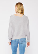 [Color: Heather Grey] A blonde model wearing a grey relaxed fit sweater with an asymmetrical neckline and long sleeves with a dropped shoulder. An everyday pullover sweater styled with denim jeans for the fall season.