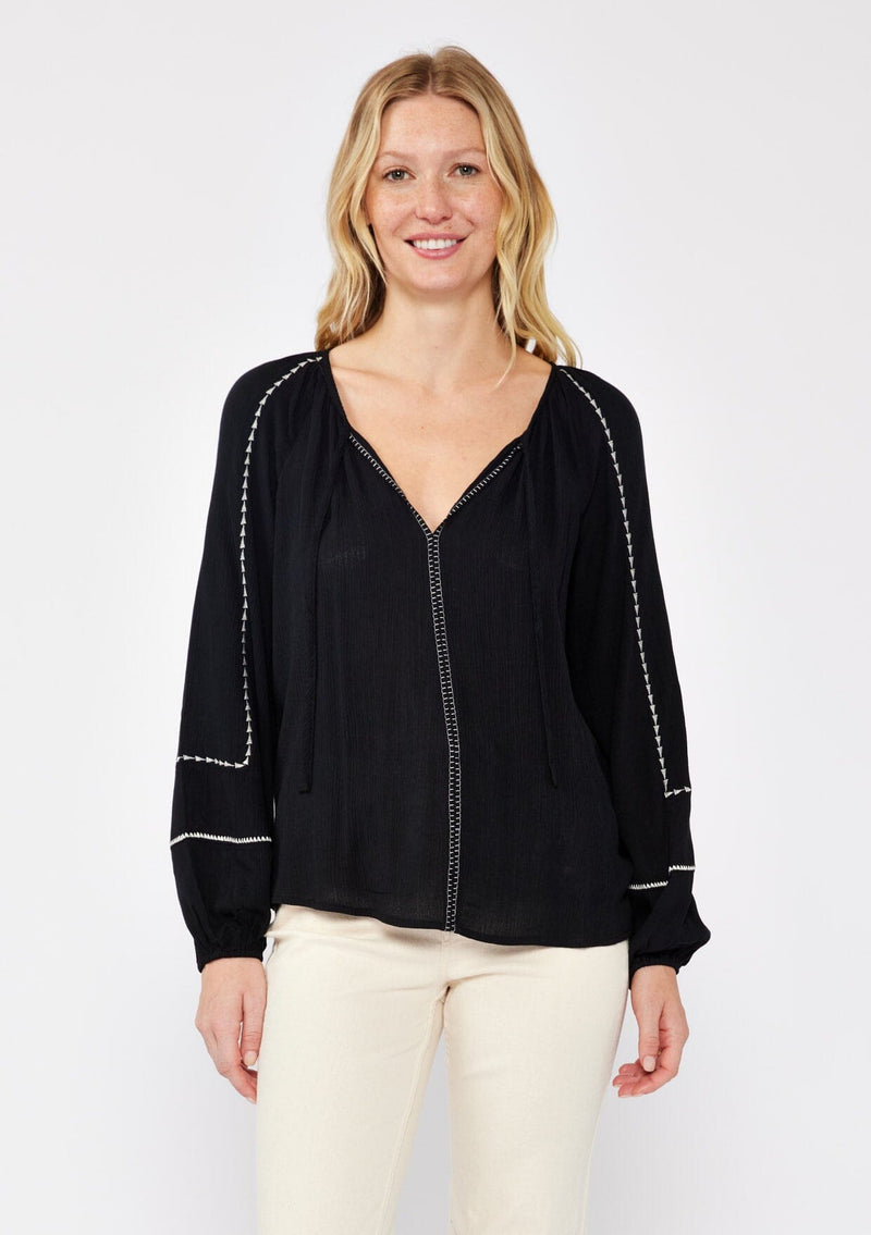 [Color: Black/Natural] A blonde model wearing an embroidered black bohemian blouse. With a split v neckline with ties, long raglan sleeves, and an elastic cuff. A relaxed fit boho top styled with beige denim for the ultimate fall ensemble. 