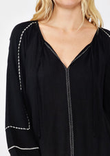 [Color: Black/Natural] A blonde model wearing an embroidered black bohemian blouse. With a split v neckline with ties, long raglan sleeves, and an elastic cuff. A relaxed fit boho top styled with beige denim for the ultimate fall ensemble.